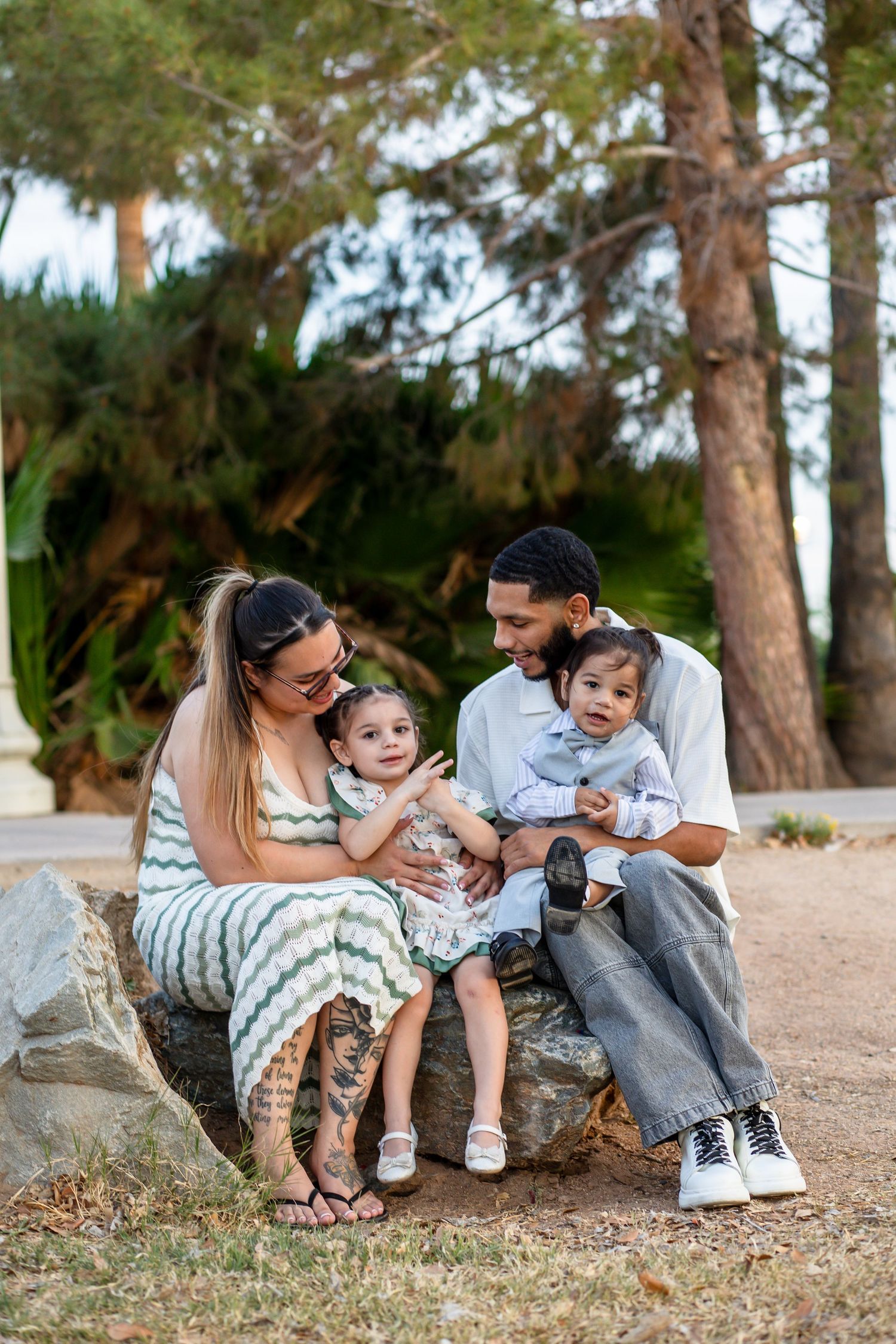 Family of four sitting on rocks in a park, parents smiling at their two young children outdoors.