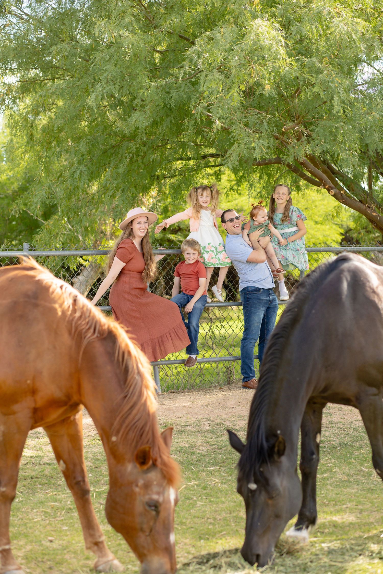Happy family posing by a fence with two grazing horses, brown and black, on a sunny farm day.