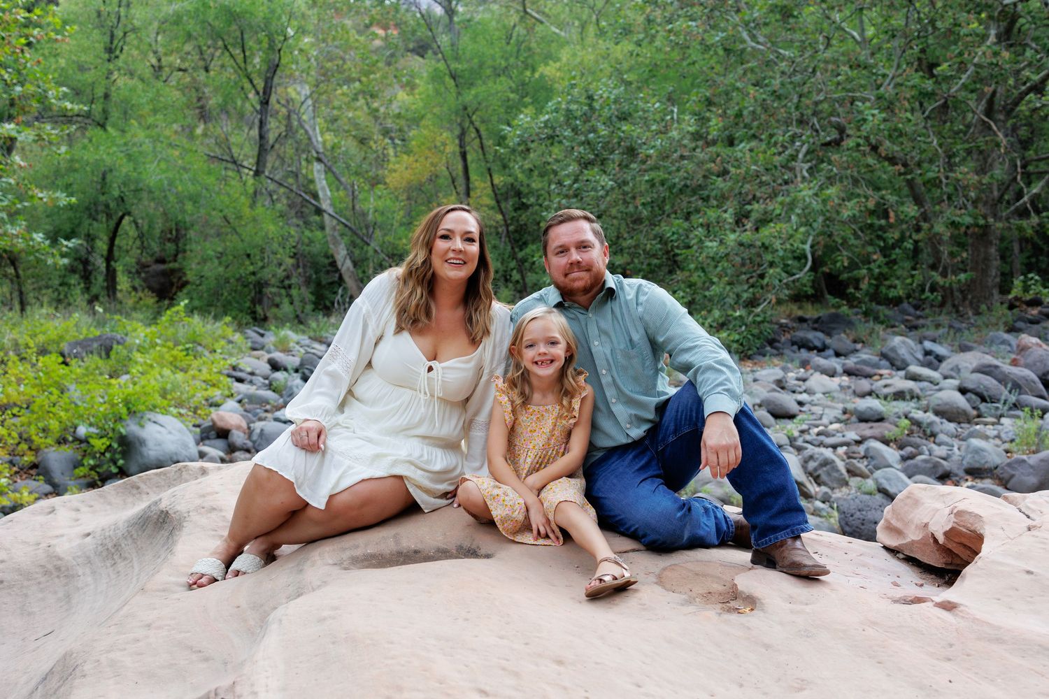 Family of three smiling on a boulder near a rocky creek surrounded by lush green trees.