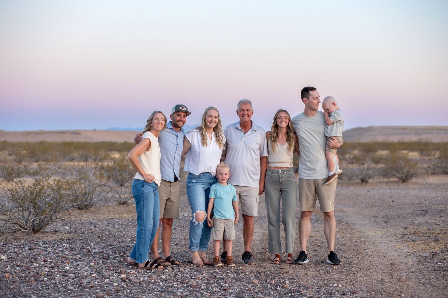 Multi-generational family smiling together outdoors in desert landscape at dusk with pink sky backdrop.