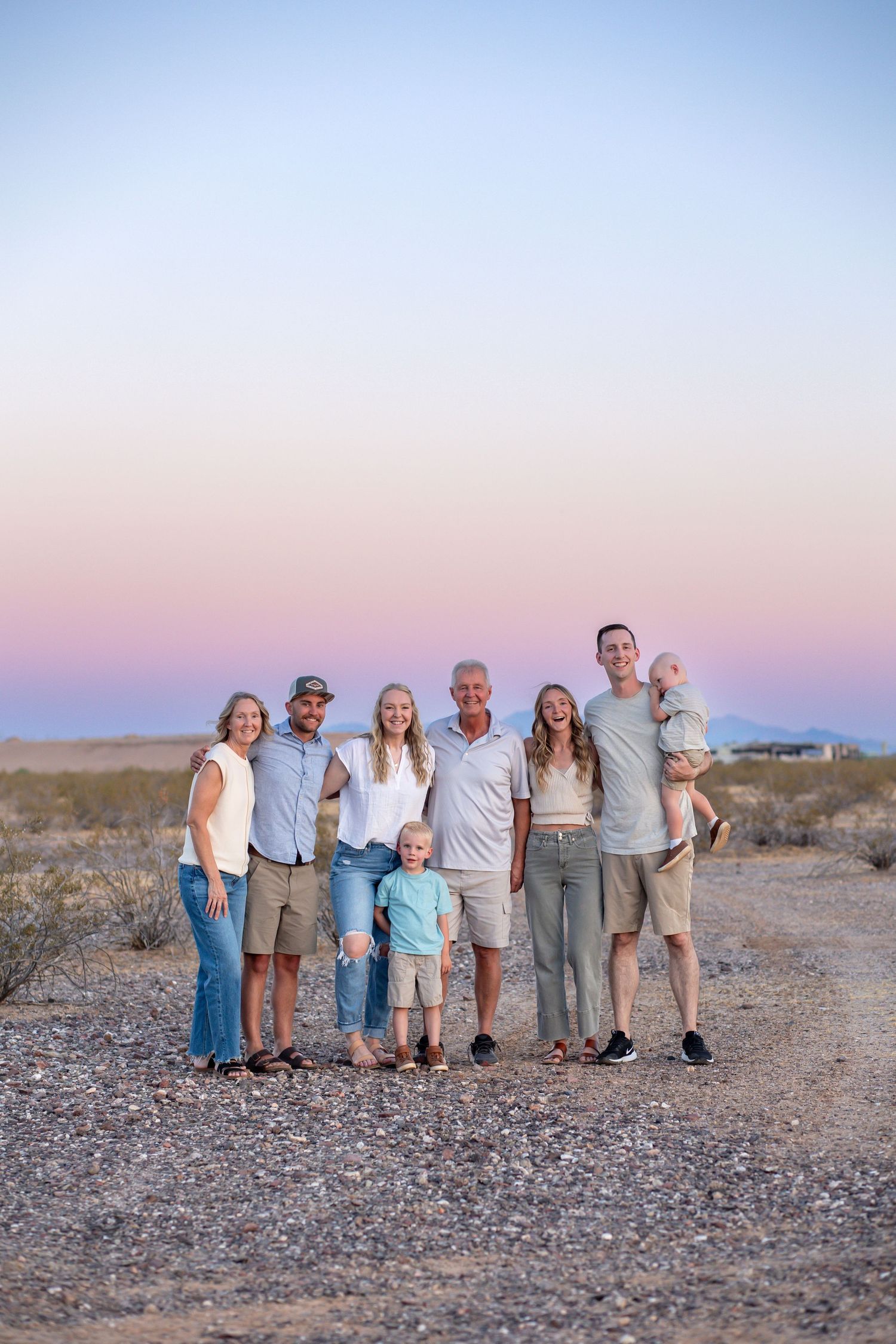 Multi-generational family posing together at desert sunset, smiling outdoors on gravel path with pastel sky backdrop.
