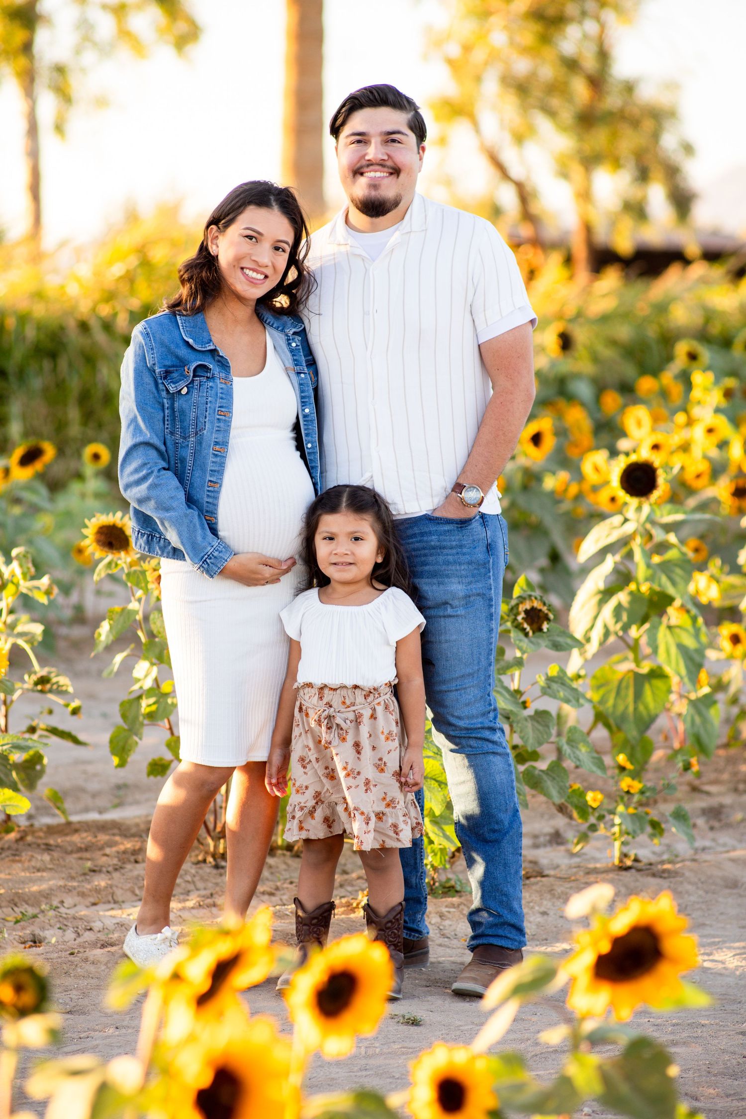 Pregnant woman with family in sunflower field at sunset, young daughter standing in front of parents.