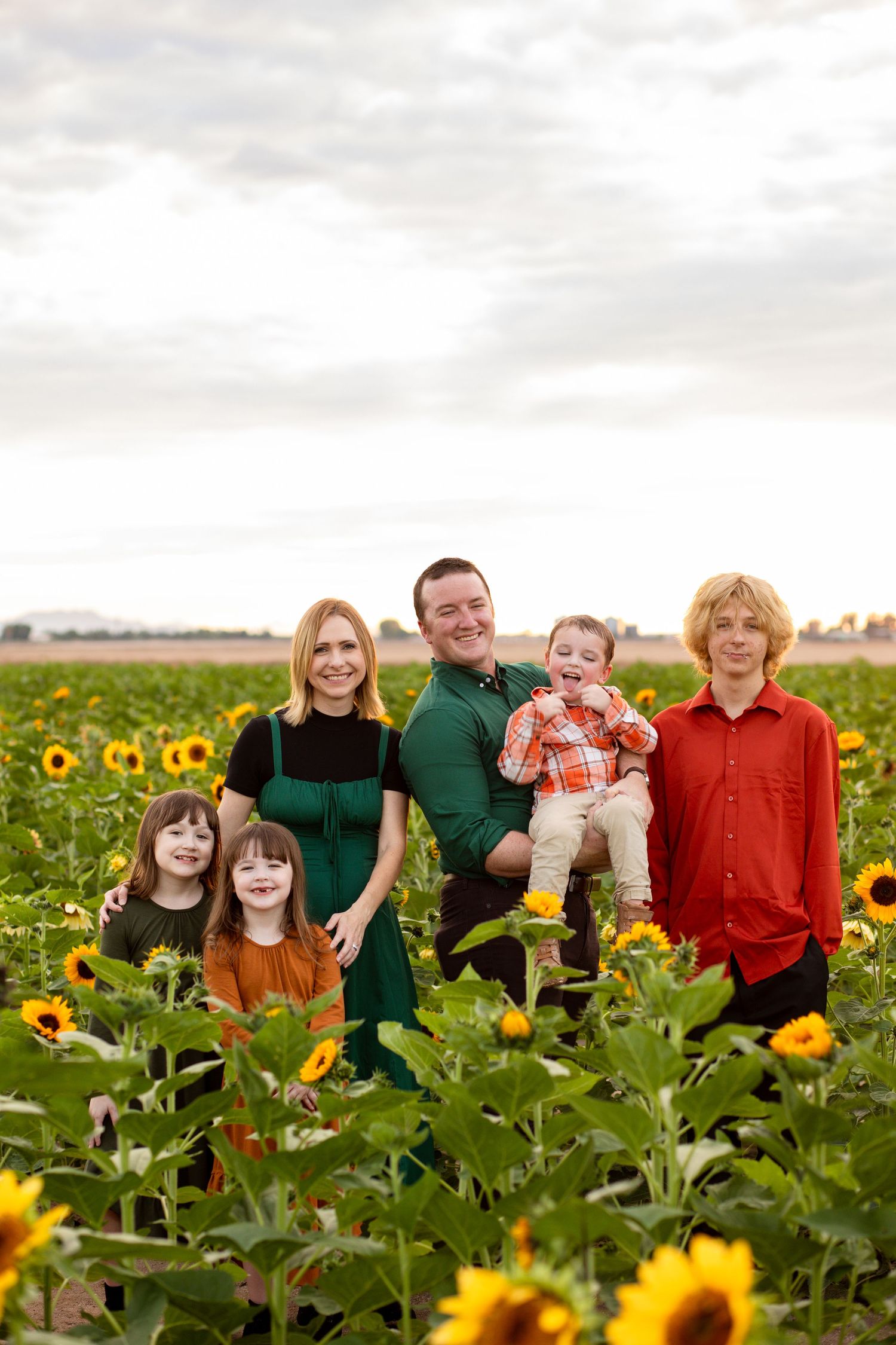 Family of six posing among vibrant sunflowers in a large sunflower field under cloudy skies.