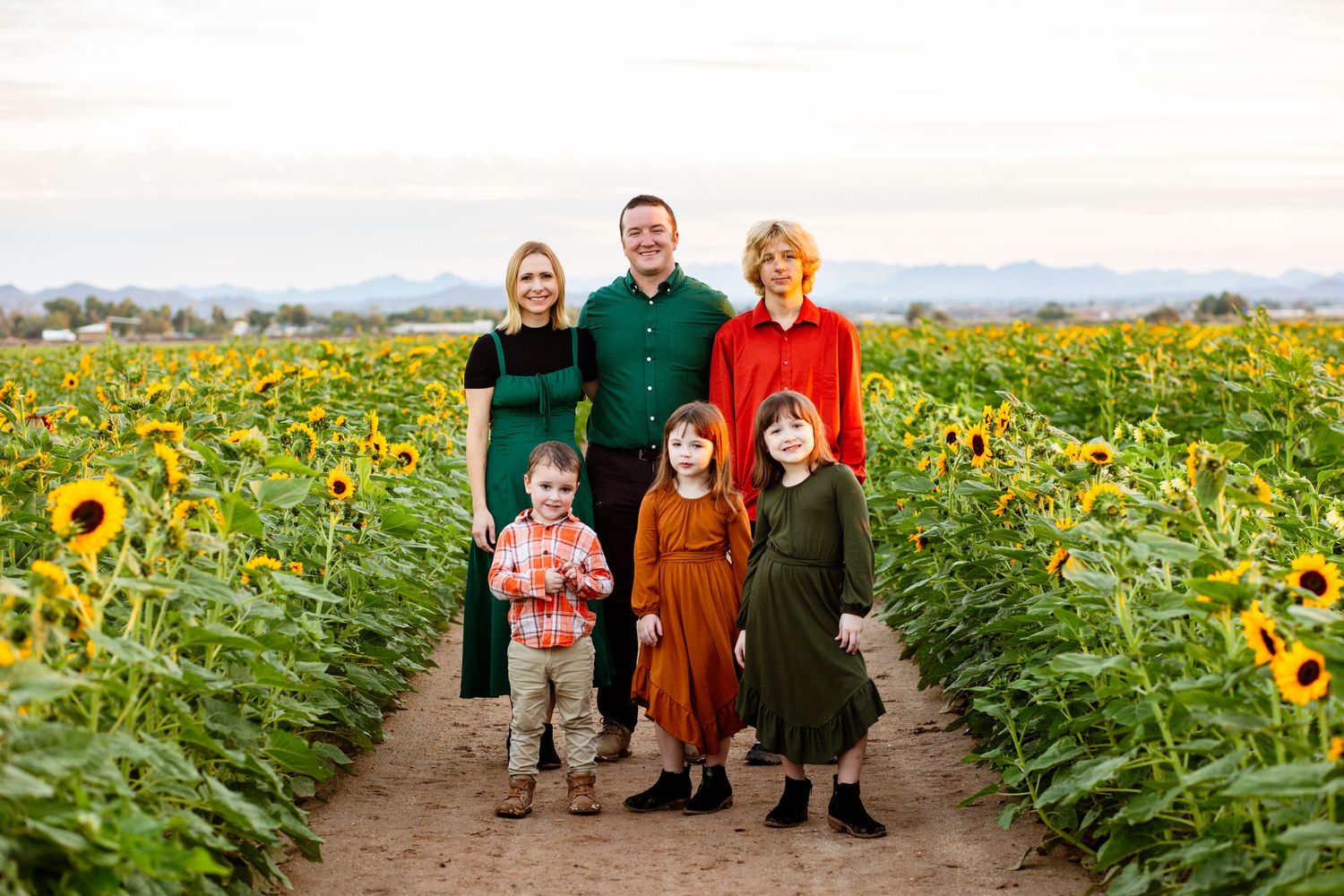 Family of six standing on path through sunflower field wearing coordinated fall colors.