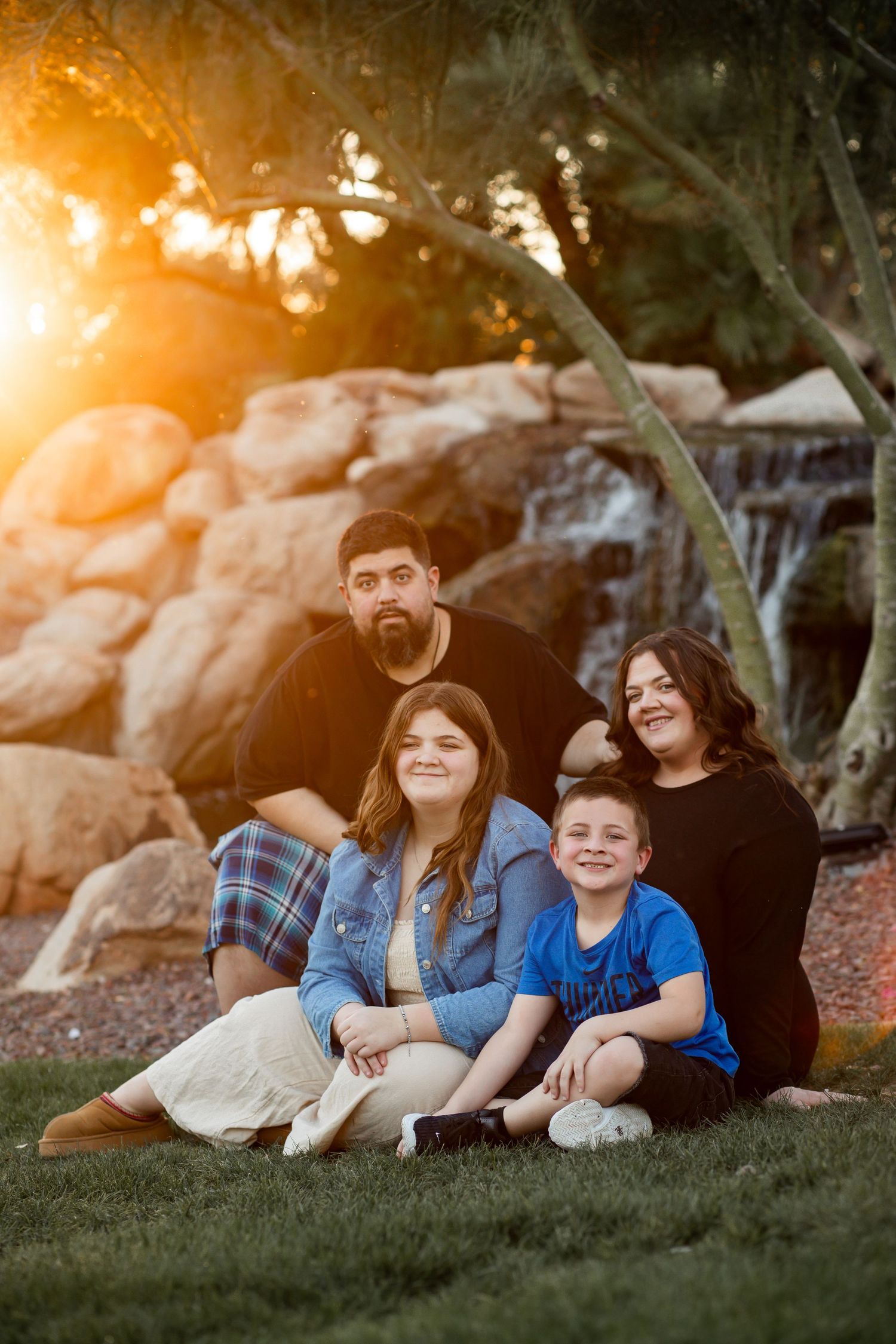Family of four sitting on grass at golden hour sunset near rocky waterfall, smiling warmly together.