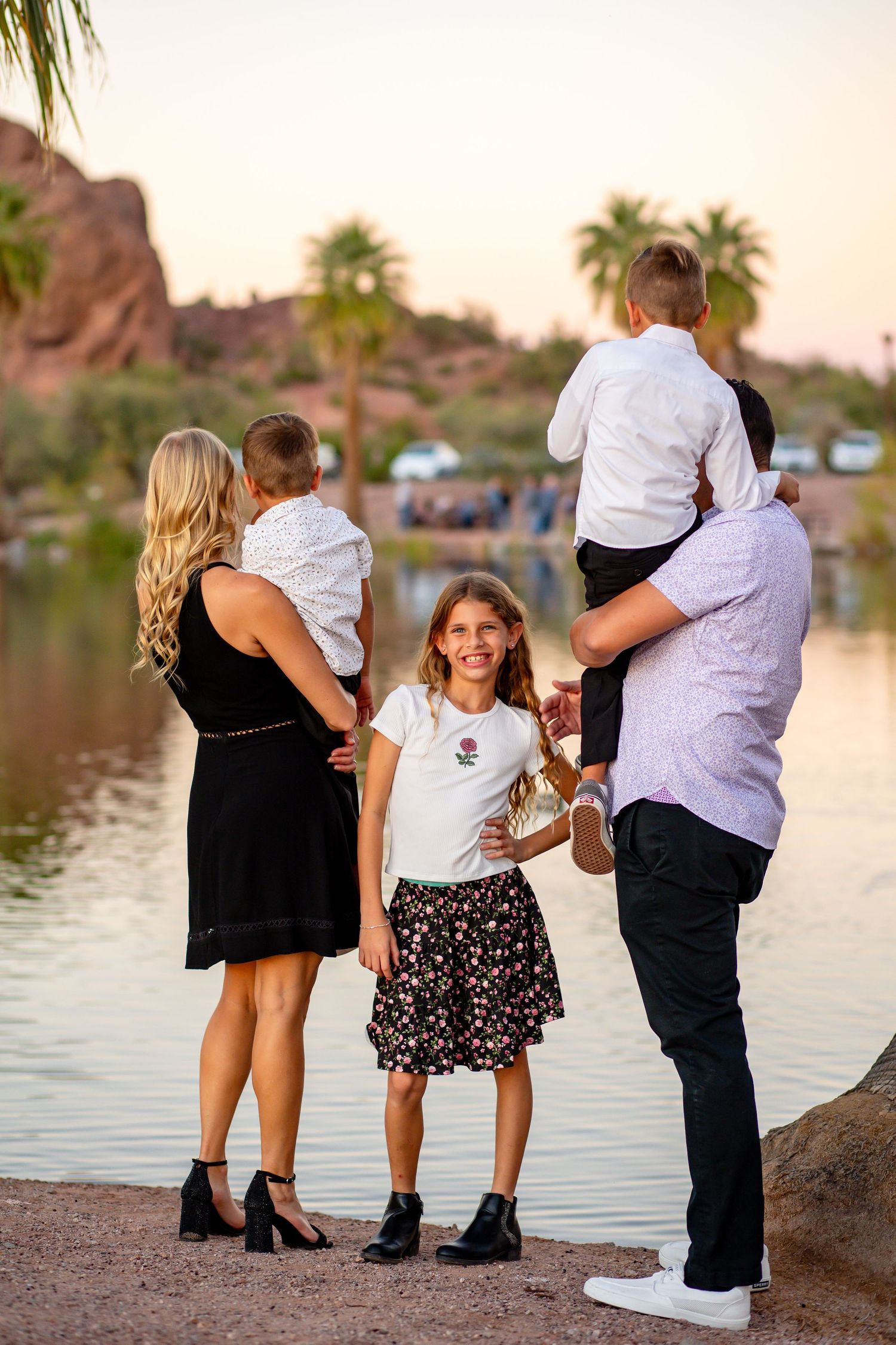 Family of five at sunset by a lake, parents holding toddlers while daughter smiles at camera.