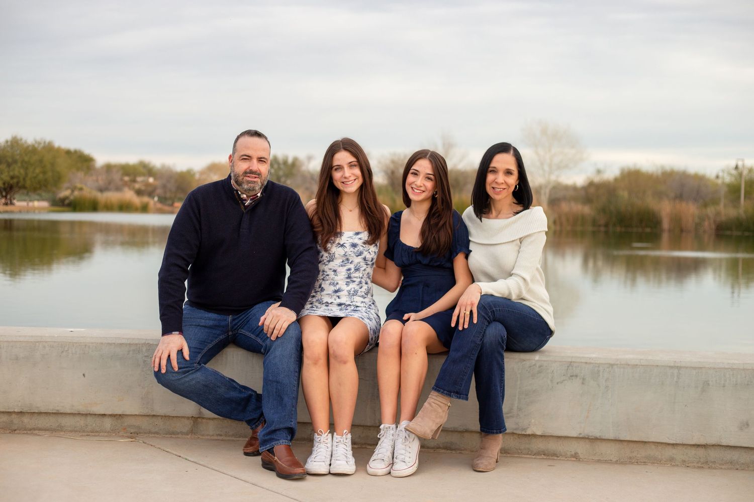 Family of four sitting by a lake, two teenage daughters between parents, all wearing casual clothing.