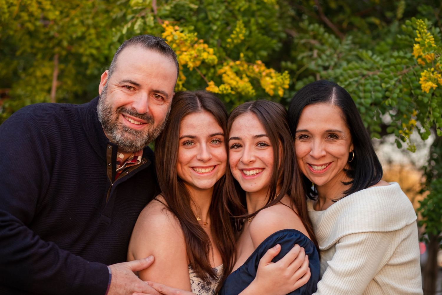Happy family of four smiling closely together outdoors surrounded by lush green and yellow foliage.