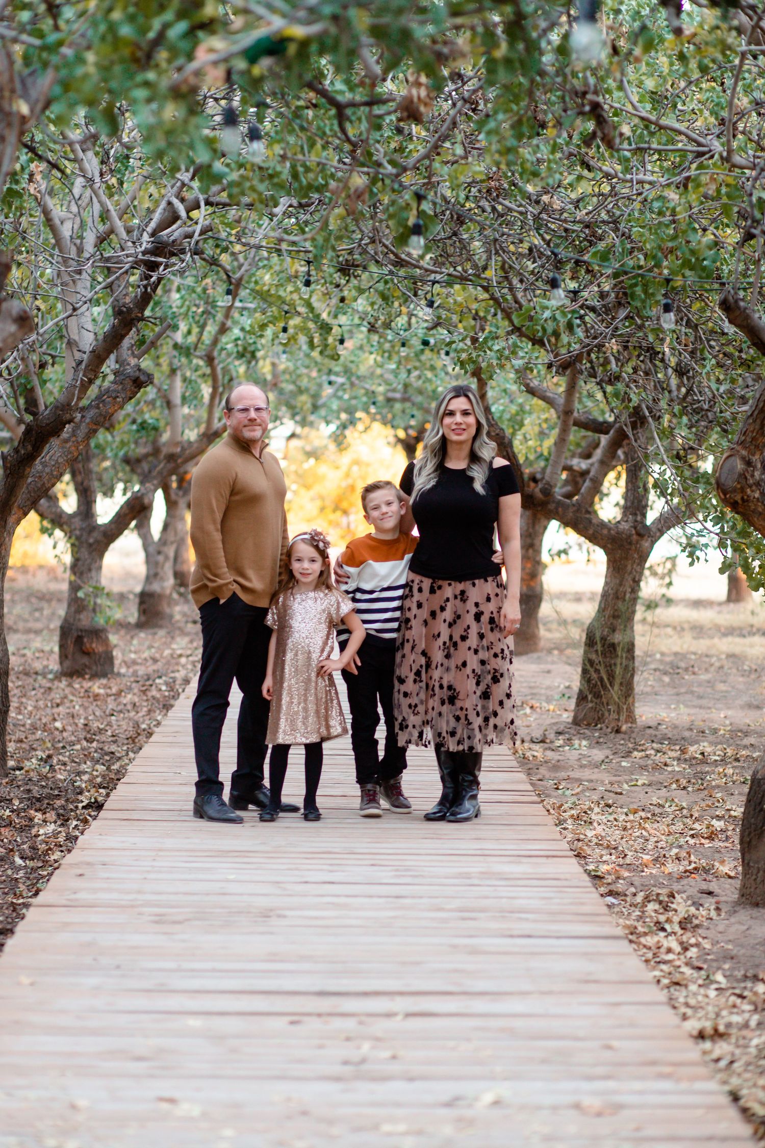 Family of four standing on wooden boardwalk path surrounded by autumn trees in park.