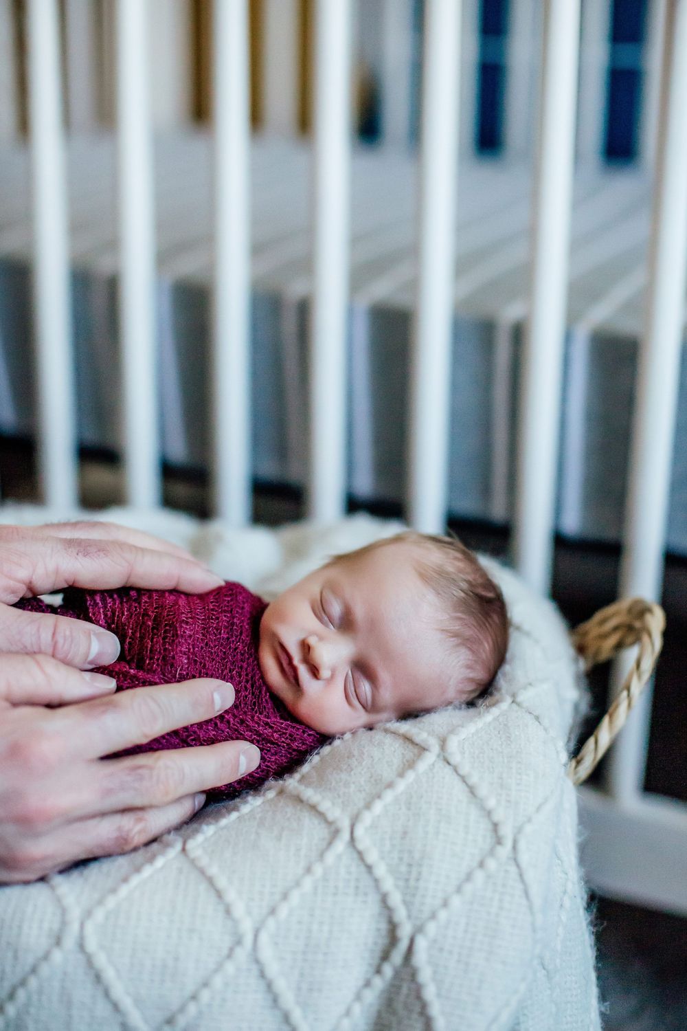 Sleeping newborn baby wearing burgundy sweater rests peacefully on white textured blanket in crib.