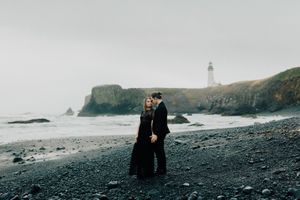 Dramatic coastal landscape with black sand beach, lighthouse, and rocky cliffs in moody lighting.
