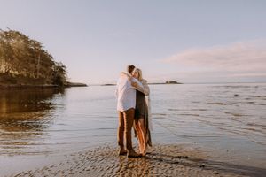 Couple walking along a scenic beach shoreline at sunset with rocky cliffs in the background.