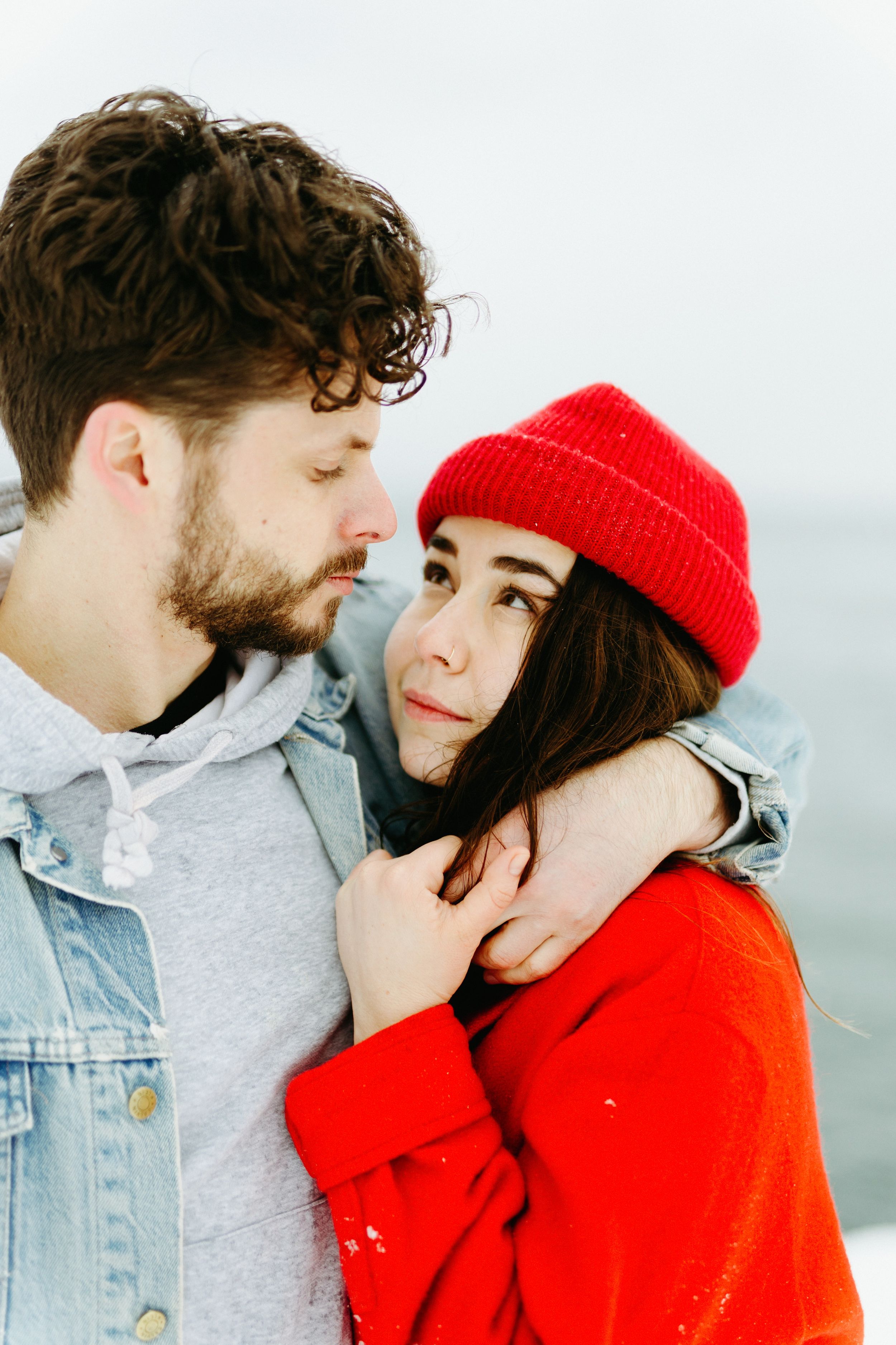 Couple embracing while one wears a bright red knit hat against a soft white background.