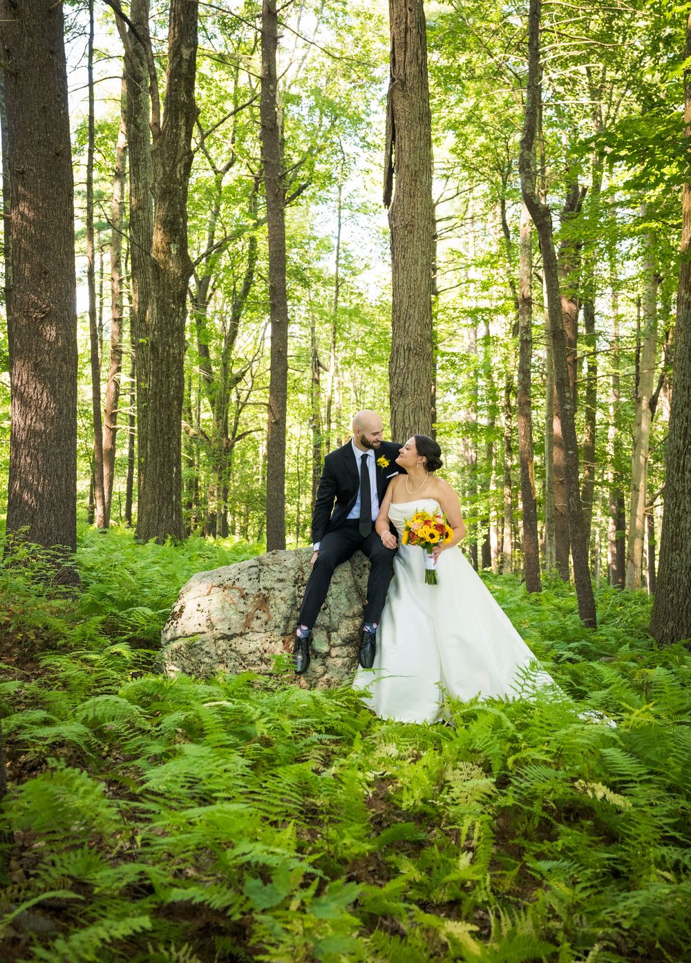 A wedding couple poses among tall trees and ferns in a magical forest setting.