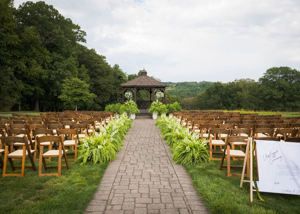 A stone pathway leads through rows of wooden chairs to a gazebo in an outdoor garden wedding ceremony setup.