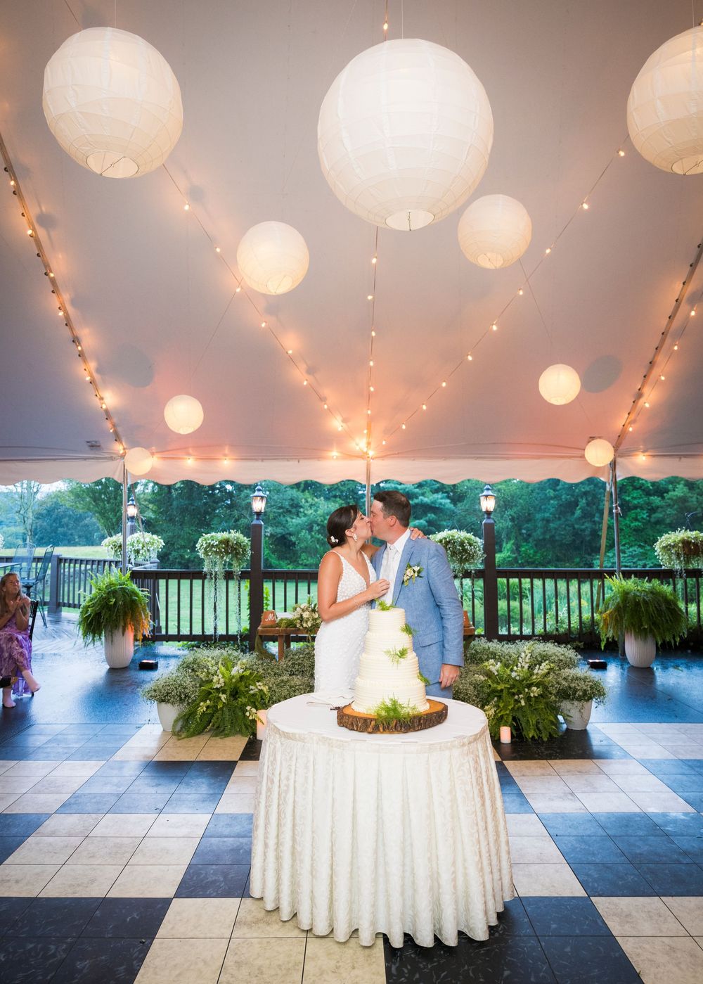 A romantic wedding scene under a tent with twinkling lights, white decor, and lush greenery overlooking a scenic view.