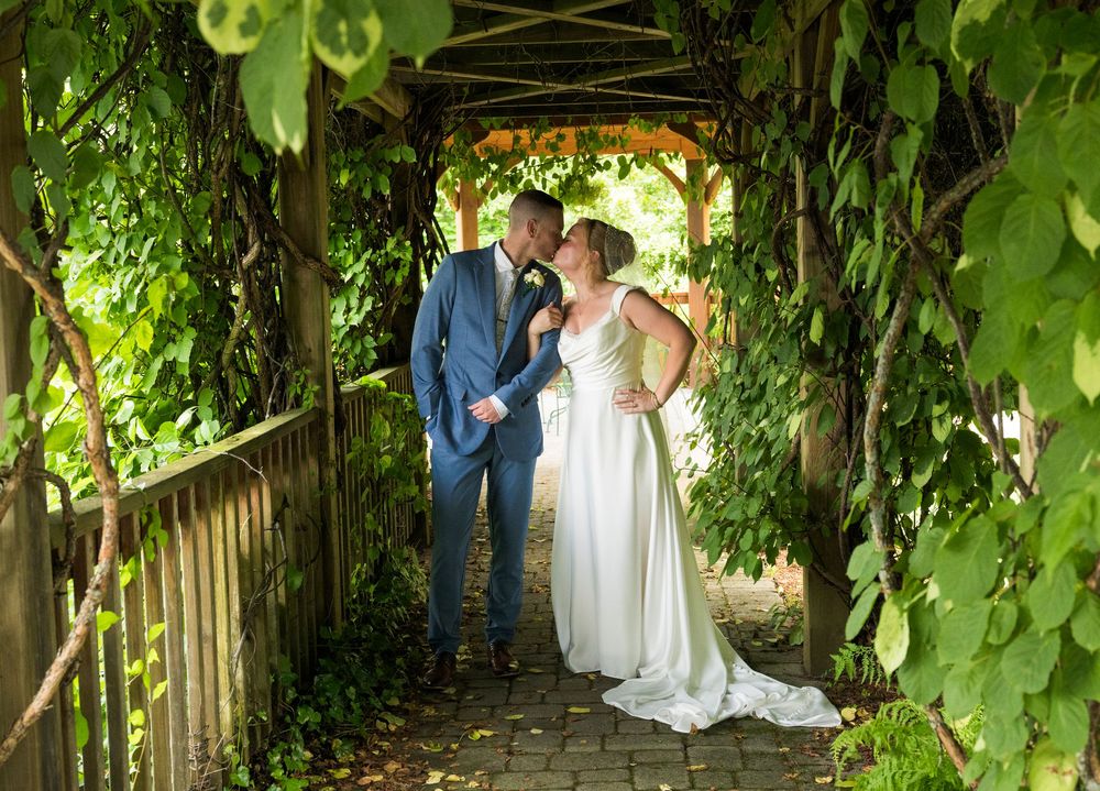 Wedding couple and guests stand beneath a lush green archway of vines and leaves during an outdoor ceremony.