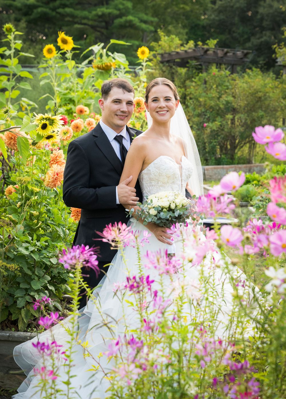 A couple poses in a garden filled with vibrant pink flowers and sunflowers on their wedding day.