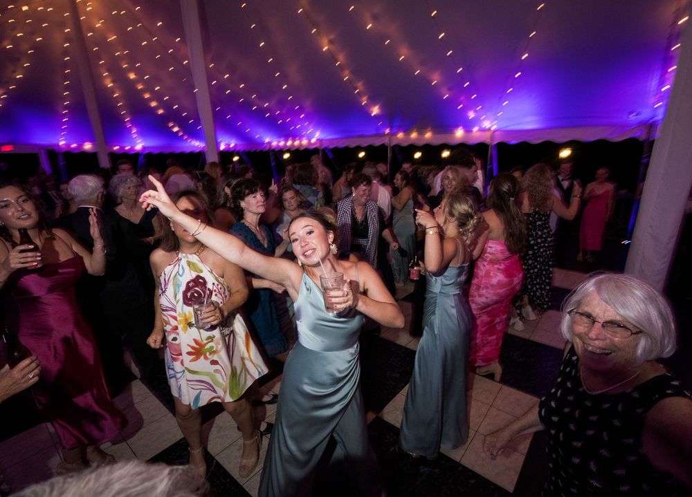 Wedding reception dance floor filled with guests celebrating under purple mood lighting and string lights.
