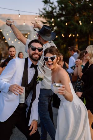 Wedding celebration with guests wearing sunglasses and dancing under outdoor string lights at dusk.