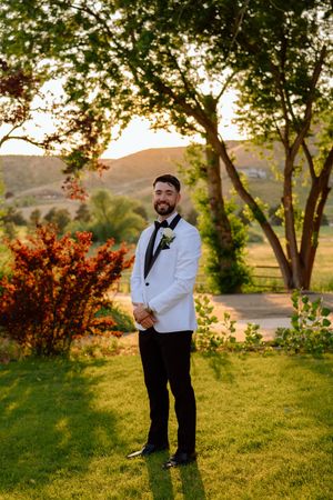 Groom in white dinner jacket poses on lawn at sunset with mountains in background.