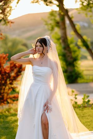 A bride in a white strapless gown with flowing veil poses in golden sunlight during sunset.