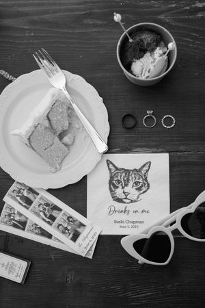 Wedding details flatlay with rings, cat illustration, and food on dark wooden surface.