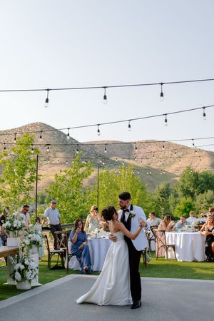 A couple dances on an outdoor patio with string lights and mountain views at their wedding reception.
