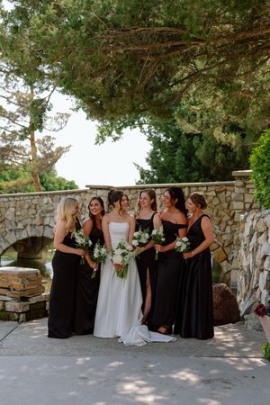 Wedding party in black and white dresses posing together against a stone wall at an outdoor venue.
