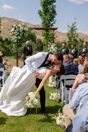 Wedding party guests jump in unison on a grassy lawn with mountains in the background during an outdoor ceremony.