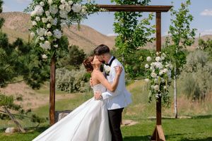 A couple shares a romantic kiss under a wooden arch decorated with white flowers in an outdoor mountain setting.