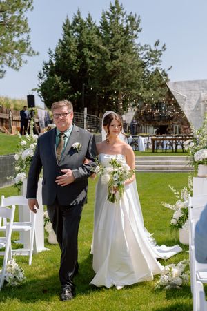 Outdoor wedding ceremony setup with white chairs and floral arrangements along the aisle on a sunny day.