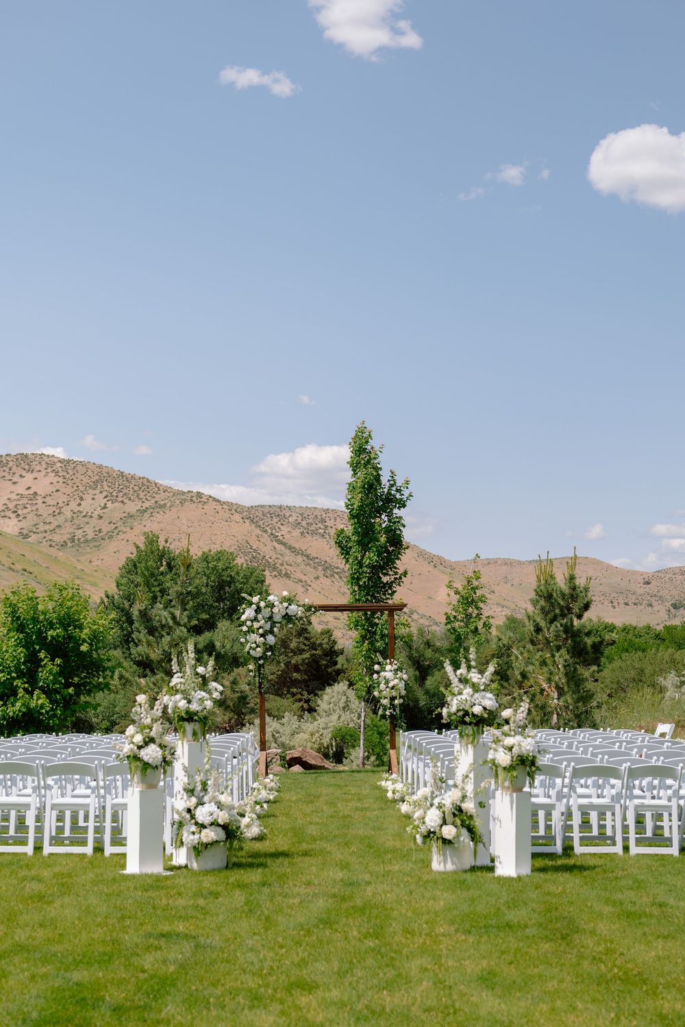White chairs and floral arrangements line a grassy outdoor wedding ceremony space with mountains in the background.