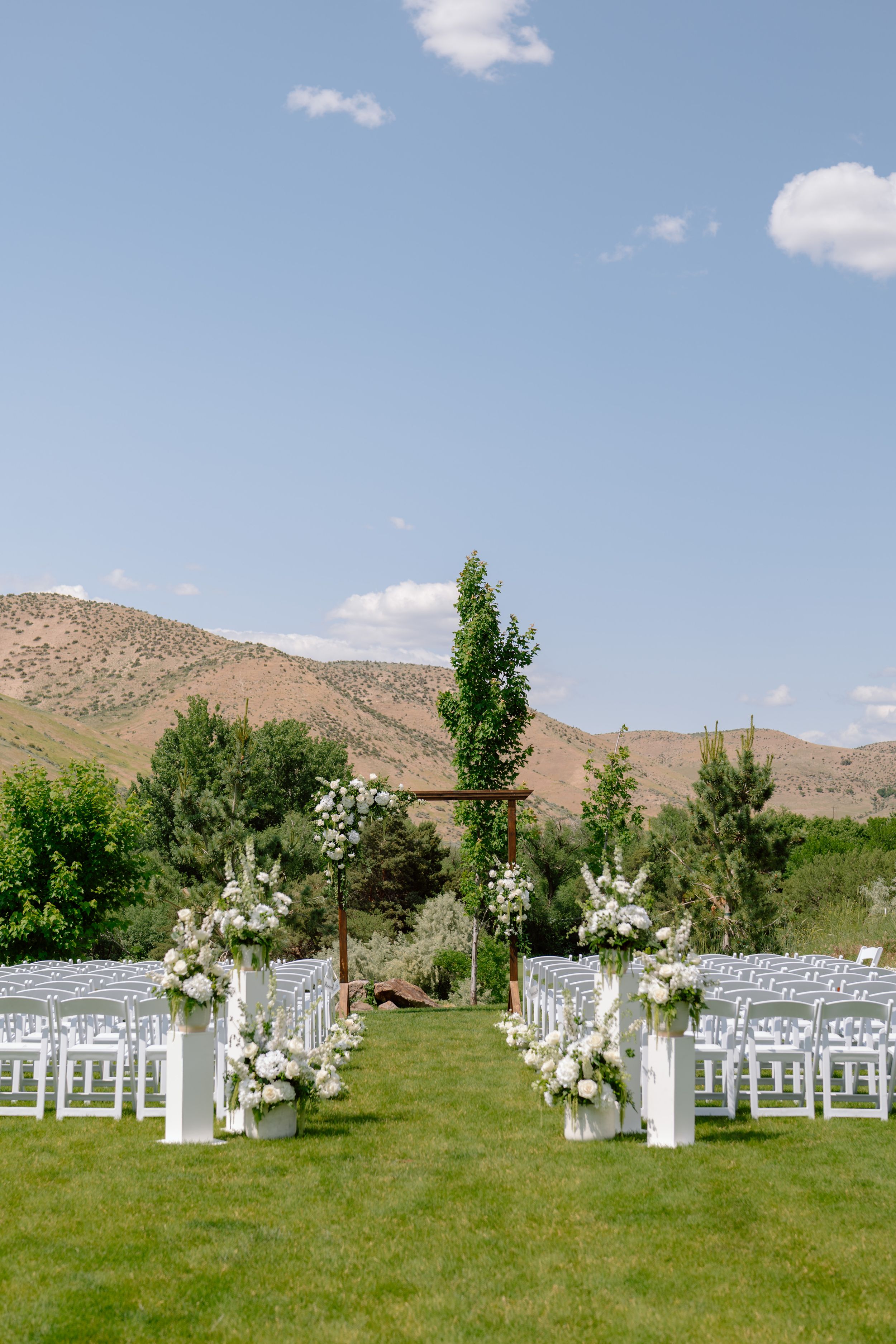 White chairs and floral arrangements line a grassy outdoor wedding ceremony space with mountains in the background.