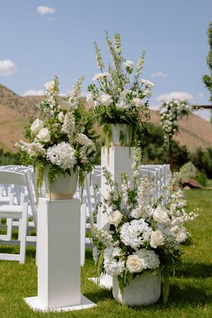 White floral arrangements in modern vases line an outdoor wedding aisle with white chairs against a mountain backdrop.