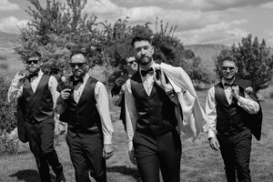 Groomsmen in black vests pose together outdoors in a playful jumping photo against a cloudy sky.