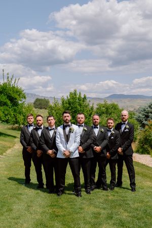 Groomsmen in black suits and ties pose together on a grassy hillside with mountains visible in the background.