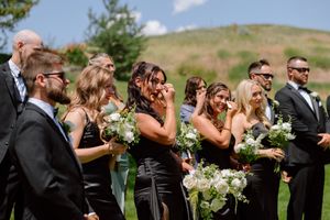 Wedding party members in formal attire pose together on grass with white floral bouquets against mountain backdrop.