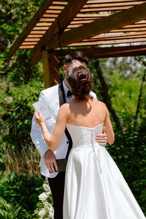 A couple shares an intimate moment under a wooden pergola during an outdoor wedding ceremony.