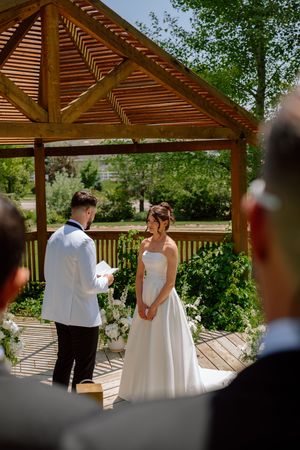 Outdoor wedding ceremony taking place under a wooden pergola surrounded by lush greenery on a sunny day.