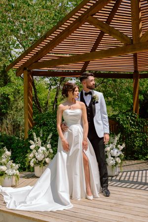 A wedding couple shares a tender moment under a wooden pergola surrounded by lush greenery and white floral arrangements.