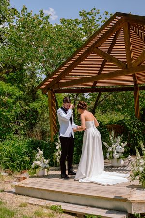 A couple stands under a wooden pergola during an intimate outdoor wedding ceremony in a lush garden setting.