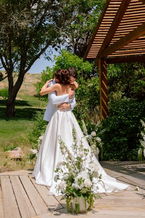 A bride in an elegant white wedding dress stands on a wooden deck under a pergola surrounded by greenery and flowers.
