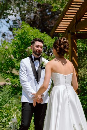 A romantic wedding moment captured outdoors under a wooden pergola with lush greenery in the background.