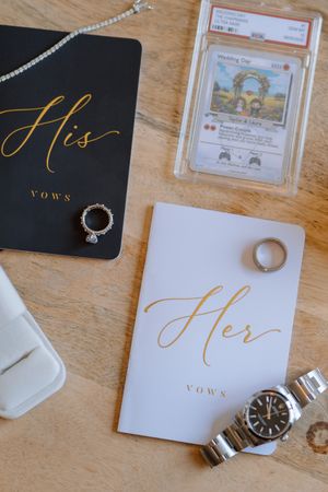 Wedding vow books in black and white with gold lettering displayed alongside rings and watches on wooden surface.
