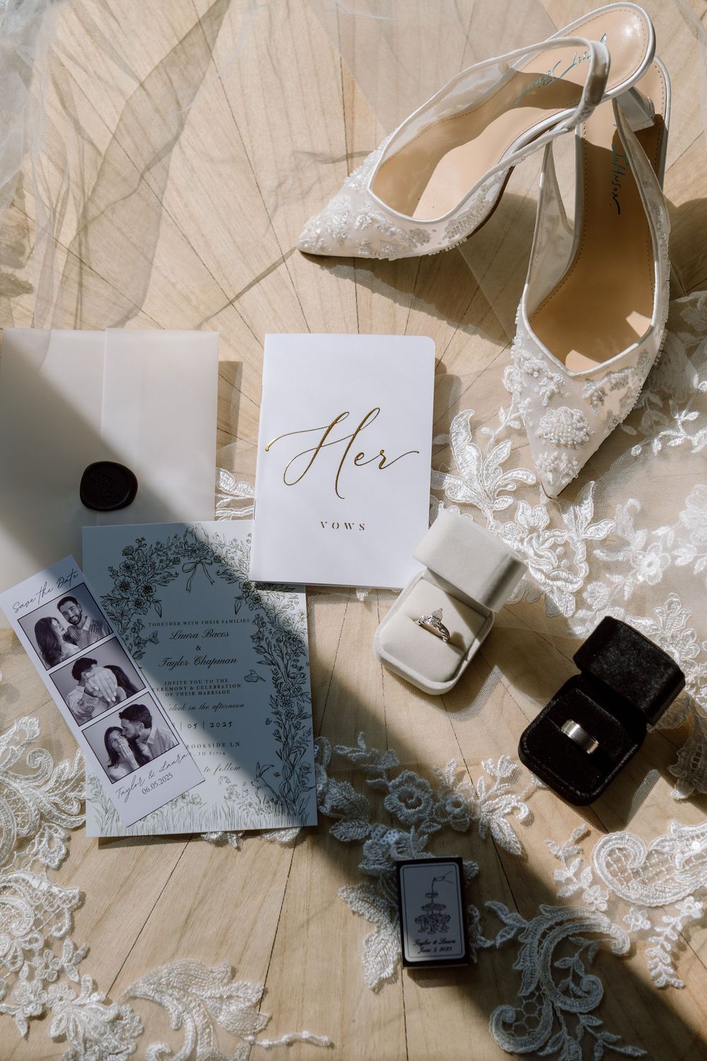 A collection of wedding details including white heels, lace, invitations and perfume bottles arranged on a light wooden surface.
