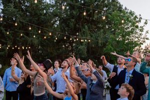 Wedding guests raise their arms and dance together under string lights at an outdoor evening celebration.