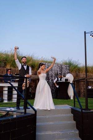 A joyful wedding couple celebrates with raised arms on outdoor steps at sunset.
