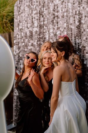 Wedding party members in black dresses pose playfully with the bride in a white gown against a textured backdrop.