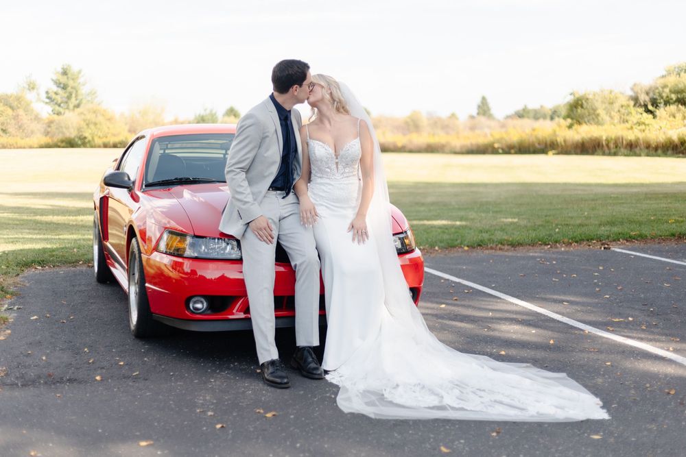 Wedding couple poses with red sports car in parking lot.